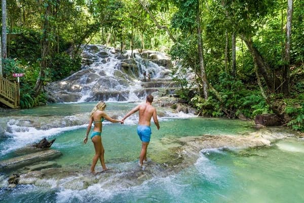 A couple walking through shallows in a lush tropical setting, silhouetted by a waterfall